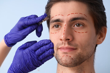 Plastic surgeon with scalpel and markings on young man's face against blue background, closeup