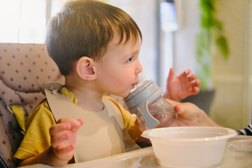 Mother woman helps baby boy drink water from a bottle of non-spill bottle in a cafe. A child eats in a restaurant sitting at a table on a high chair. Kid aged two years - Powered by Adobe