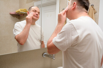 A man wipes his face with a cotton pad in the bathroom in front of the mirror. Overweight...