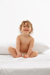 Obraz na płótnie Canvas Cheerful infant in playful pose, sitting up on white mattress with bright smile, fun expression against white background. Concept of beauty, childhood, motherhood, life, birth. Copy space for ad