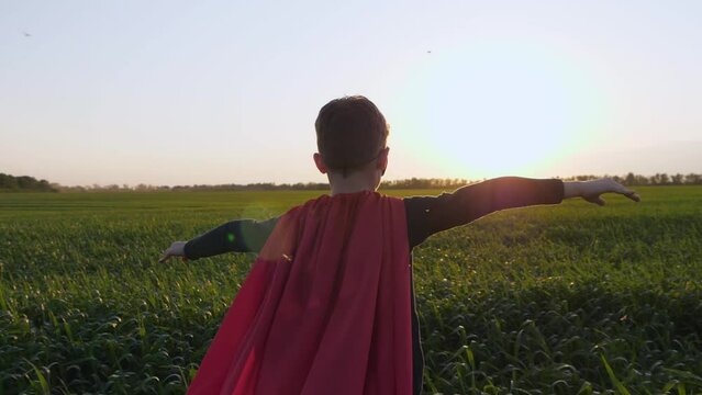 A child in a red cape portrays a superhero. boy superhero back view running through a green meadow to meet the sunset. arms outstretched to the side in flight.