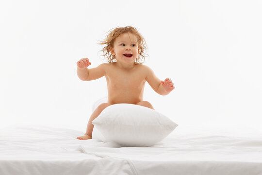 Playful and funny little baby-girl playing, have fun on bed and jumping on soft pillow against white background. Concept of childhood and carefree, motherhood, life, birth. Copy space for ad