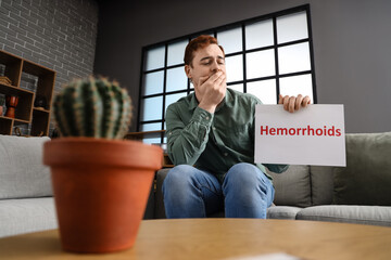 Young man holding paper with word HEMORRHOIDS on sofa at home