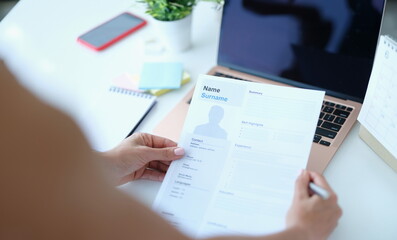 Woman sitting at table in front of laptop and holding paper with resume for employment in her...
