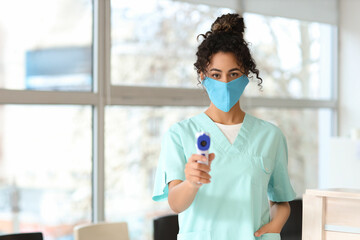 Female African-American medical intern in mask with infrared thermometer at hospital
