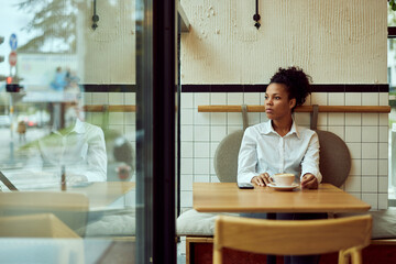 Side profile of an African woman, sitting by herself at the cafe, drinking a cup of coffee, looking...