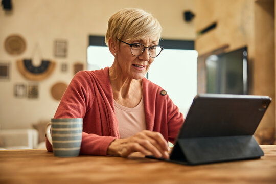 A senior woman sitting at home and using a digital tablet while drinking a cup of coffee.