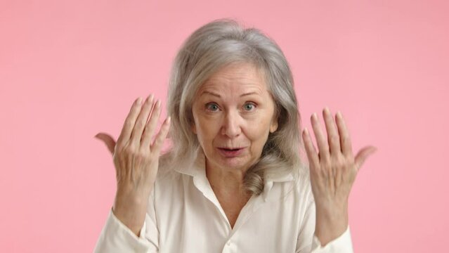An elderly woman's face conveys strong disapproval and dissatisfaction as she gestures emphatically in a video, with a soft pink background underscoring the tension. High quality 4k footage
