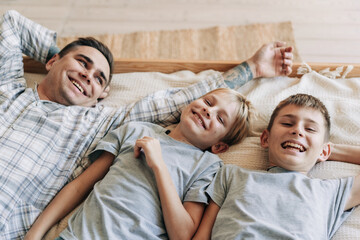 Happy laughing dad and sons are fooling around and having fun while lying on the bed.