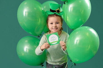 Fototapeta na wymiar Cute little girl with leprechaun's hat, greeting card and balloons on green background. St. Patrick's Day celebration