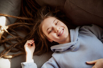 View from above. Portrait of smiling young lady with long dreadlocks in blue hoodie lying on couch and looking at camera. Concept of hobby, lifestyle, leisure time, self-expression, fashion and style.