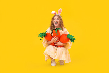 Beautiful young shocked woman in bunny ears with carrot-shaped toys on yellow background. Easter celebration