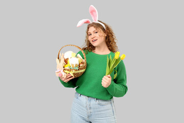 Beautiful young happy woman in bunny ears with Easter basket and tulip flowers on grey background