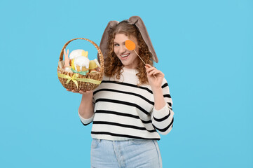 Beautiful young happy woman in bunny ears with Easter basket and paper egg on blue background