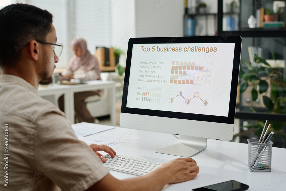 Poster Serious young businessman organizing work while sitting by desk in front of computer monitor with top five business challenges on screen