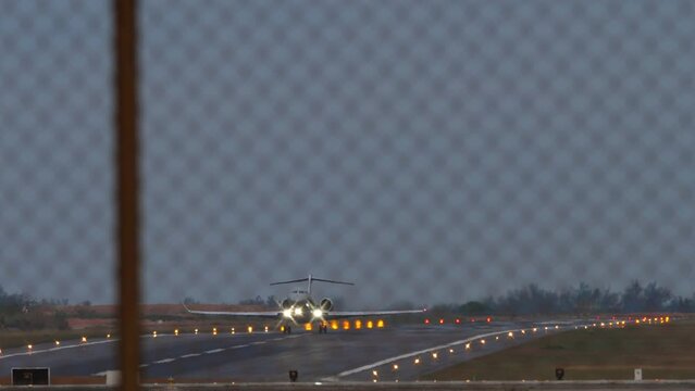 Private jet with an unrecognizable livery taking off at dusk. View through the airport fence. Landing lights, lighting, runway