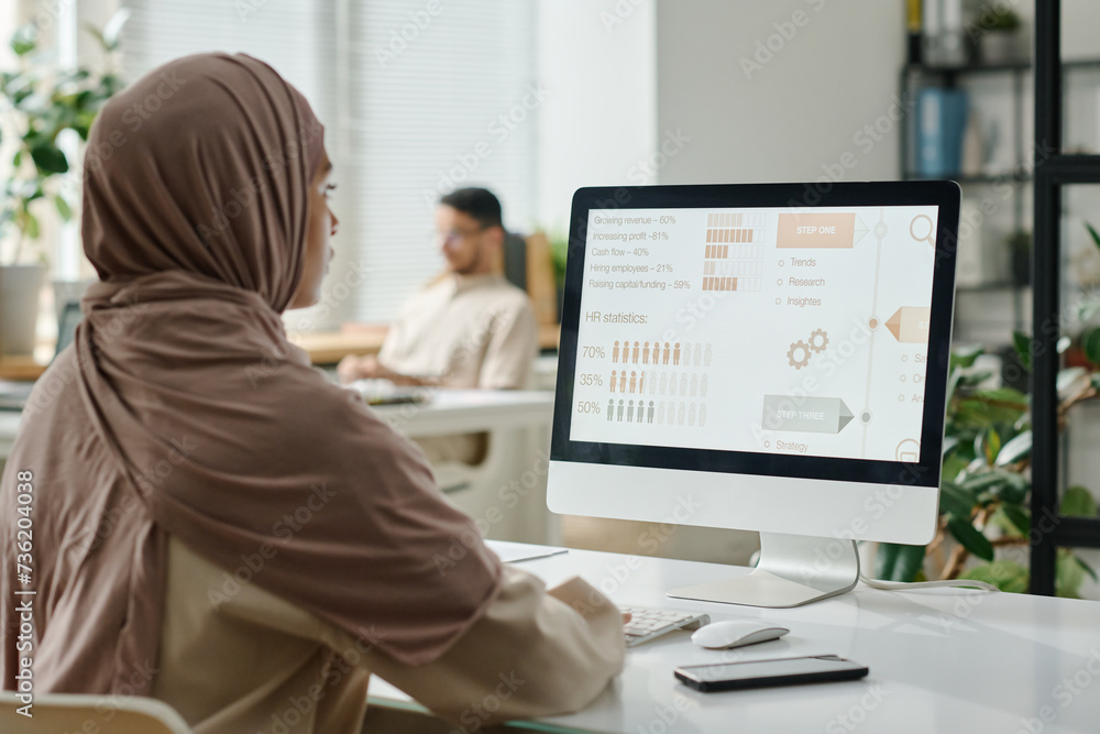 Poster young muslim businesswoman in hijab sitting by desk in front of computer screen with statistic data 