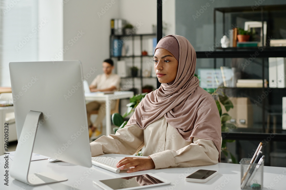 Poster young female office worker in hijab sitting by workplace in front of computer screen and analyzing d