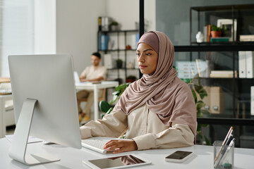 Young female office worker in hijab sitting by workplace in front of computer screen and analyzing data in coworking space