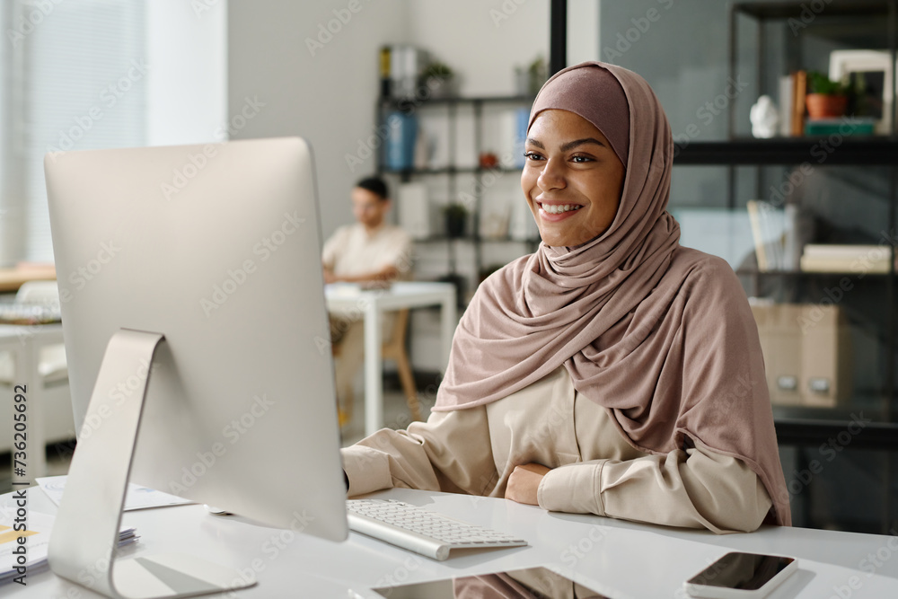 Poster happy young female analyst or broker in hijab looking at computer screen with smile while sitting by