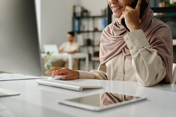 Cropped shot of young smiling office manager in hijab talking on mobile phone and clicking mouse...