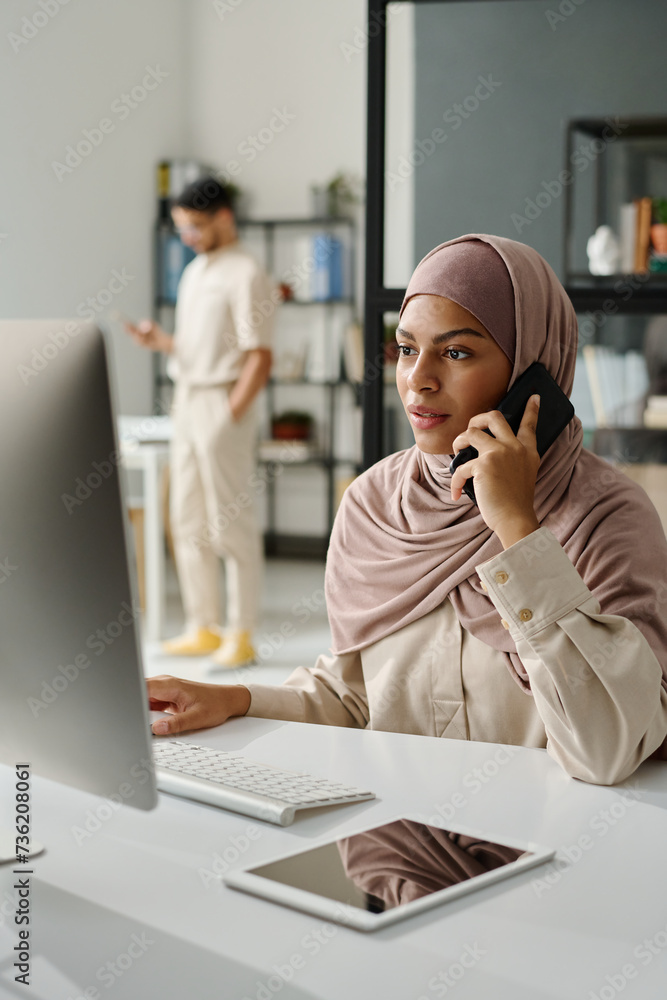 Leinwandbilder Young confident manager in hijab speaking on smartphone in front of desktop computer screen while sitting by workplace in office