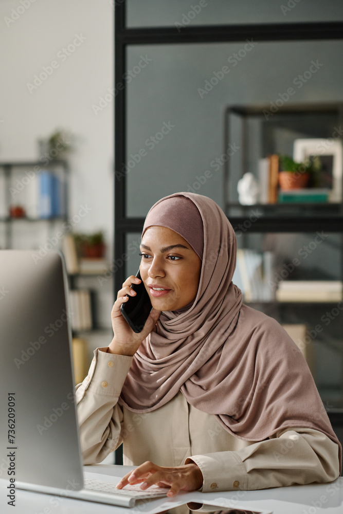 Poster muslim female manager in hijab holding smartphone by ear while sitting in front of desktop computer 