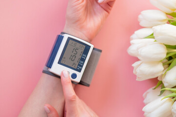 Female hands and a blood pressure monitor with flowers on a pink background.