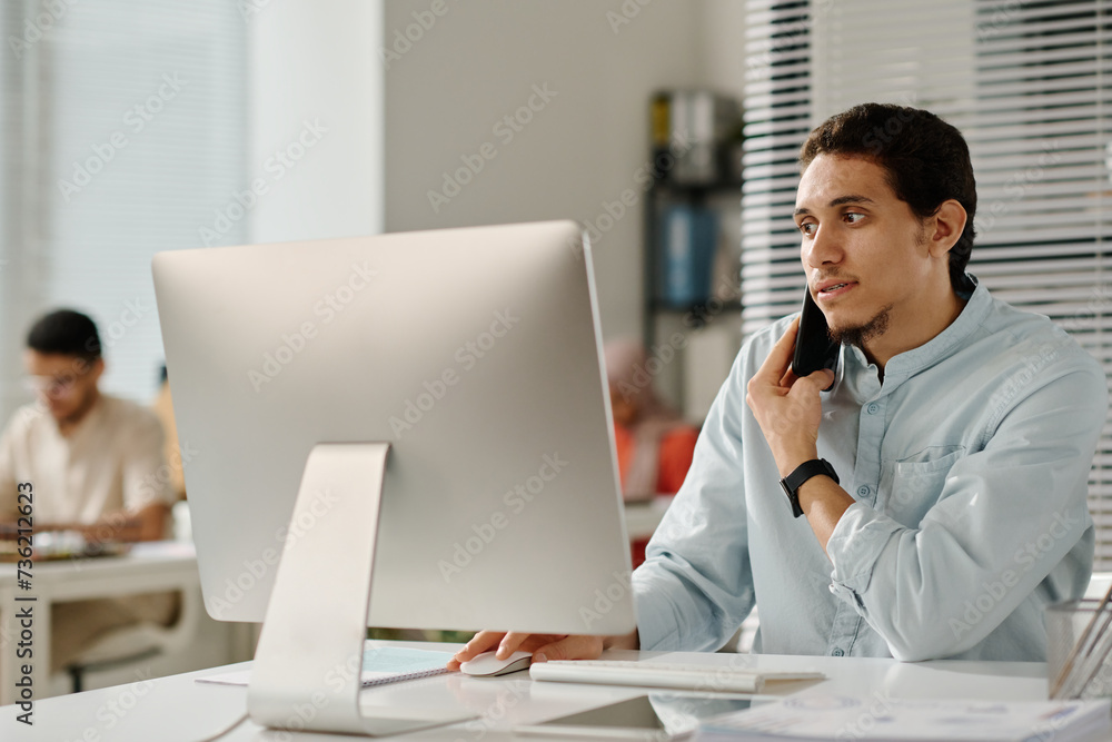 Aufkleber young confident male manager holding smartphone by ear while sitting in front of desktop computer in