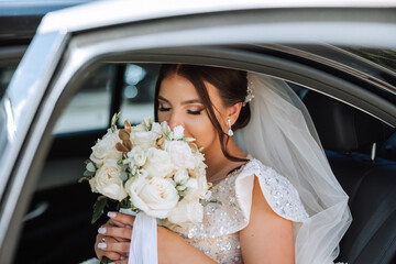 The bride is sitting in a black car with a bouquet of flowers in her hands and looks out of the...