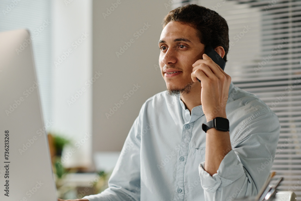 Poster young male employee talking to someone by mobile phone while sitting in front of desktop computer an