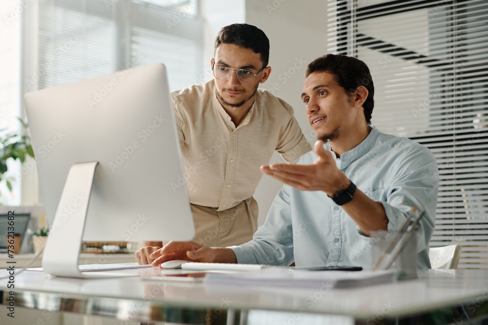 Poster two young muslim businessmen looking at screen of desktop computer while one of them explaining onli