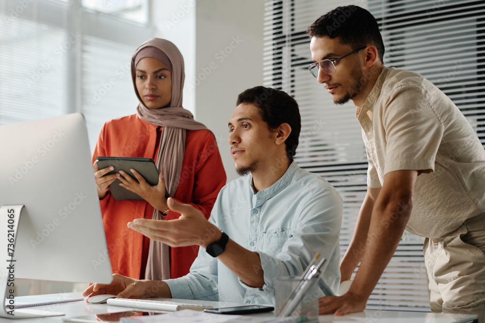 Poster team of three young muslim employees looking at computer screen while confident man explaining onlin