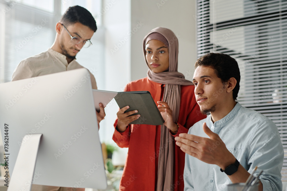 Poster two young muslim employees using tablets while standing by their colleague looking at computer scree