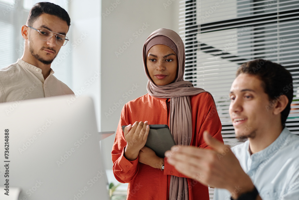 Aufkleber Young businesswoman in hijab with tablet in hands looking at computer screen and listening to explanation of data of male colleague