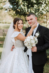 Stylish, young groom and beautiful bride in a long white dress and a long veil with a bouquet in their hands, hugging in the park in the autumn nature. Wedding portrait of newlyweds.