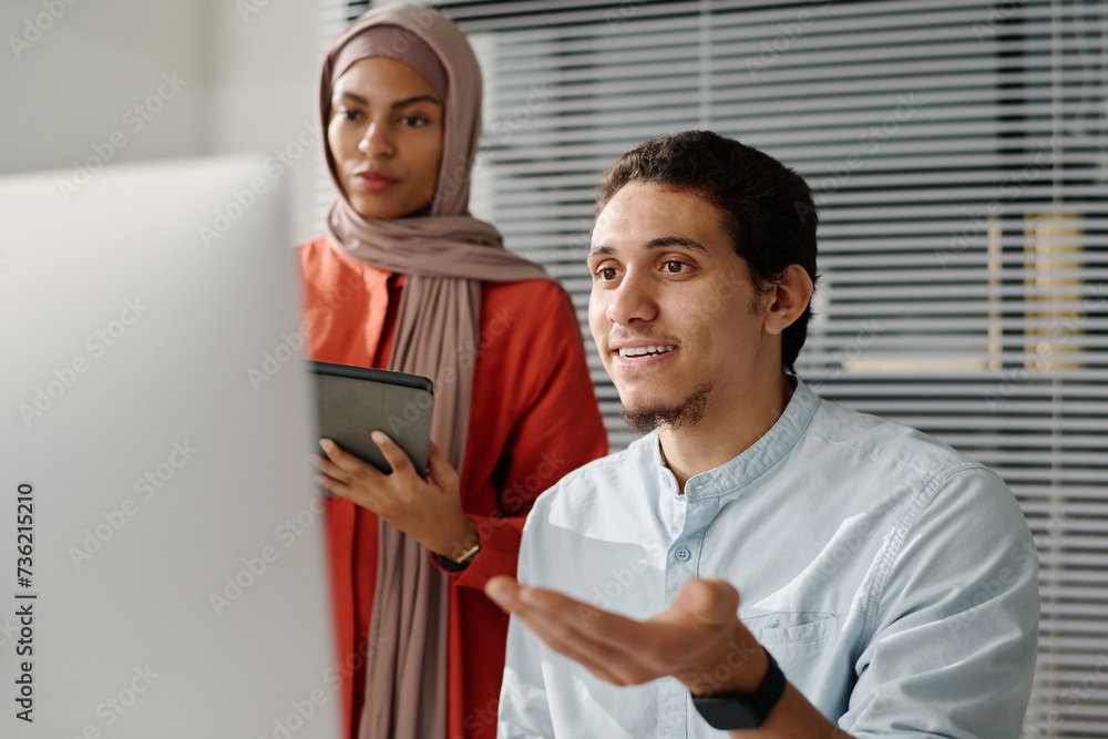 Poster young confident muslim male analyst explaining data on computer screen to female colleague in hijab 
