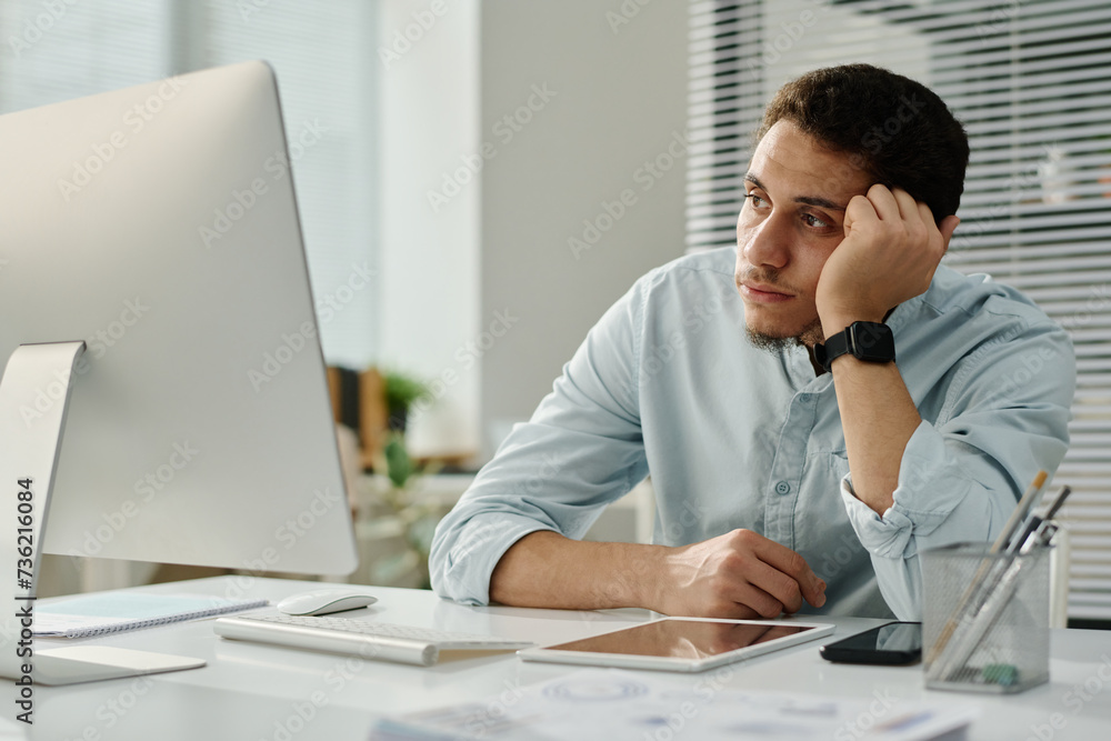 Leinwandbilder Young bored businessman sitting by workplace in front of desktop computer and looking at screen while waiting for page uploading