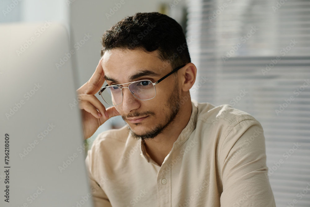 Poster young muslim male white collar worker in eyeglasses looking at computer screen while analyzing onlin