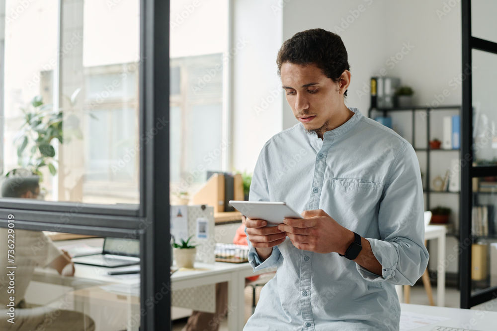 Poster young serious male analyst or broker with tablet looking through online data while standing in front
