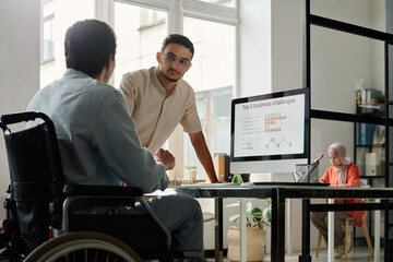 Young serious male office manager looking at his colleague in wheelchair during discussion of...