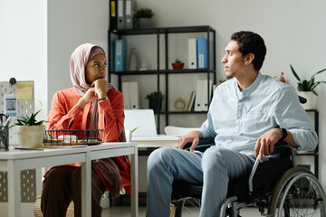 Young Muslim male office manager with disability sitting in wheelchair and looking at female...