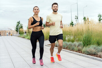 Jogging couple smiling and running together on a city walkway, with tall grass and modern...
