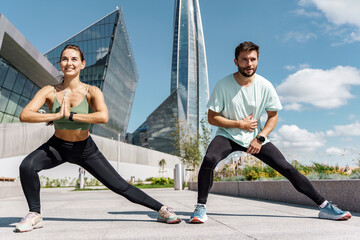 Two athletes stretching in a lunge on a sunny city plaza, with modern architecture in the backdrop.