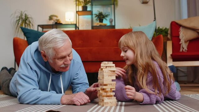 Excited little cute girl granddaughter playing building block game with smiling senior grandfather giving high five. Building wooden tower and taking out blocks in living room at home during weekends.