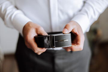 Close-up of a man holding a black leather belt. A man in a white shirt, without a face, holds a black leather belt in his hands. The morning of the groom at the wedding.