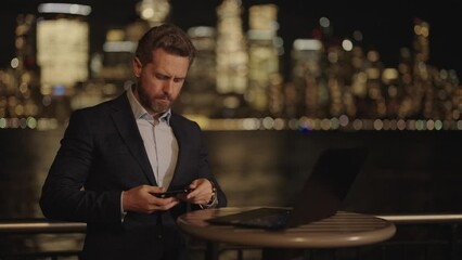 Businessman in suit using laptop talking on phone outdoor in night New York City. Night Business Life. Business man near Manhattan View on Street with Skyscrapers at Night with Neon Lights.