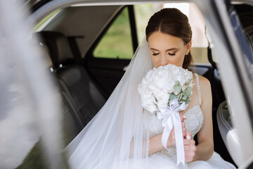 The bride is sitting in a black car with a bouquet of flowers in her hands and looks out of the car. Portrait of a rather shy bride in a car. Bride, smile, emotions.