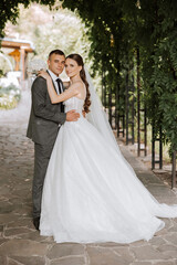 Stylish, young groom and beautiful bride in a long white dress and a long veil with a bouquet in their hands, hugging in the park in the autumn nature. Wedding portrait of newlyweds.