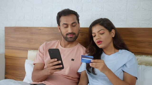 Happy romantic Indian couples shopping online using credit card on mobile phone at bedroom - concept of ecommerce purchase, relationship bonding and togetherness